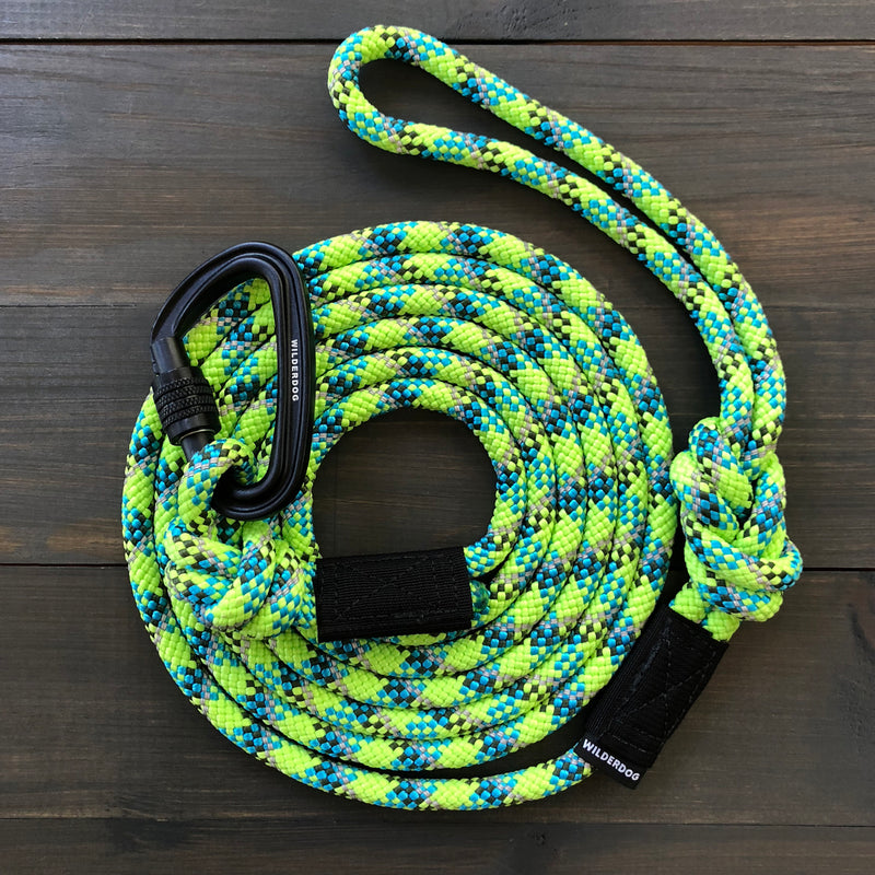 The Lime Reflective Leash, a coiled and multicolored climbing rope leash with a vibrant mix of bright green, blue, and black patterns, is laid out on a dark wooden surface. This durable dog leash features a black carabiner clip attached and a loop handle for easy holding.
